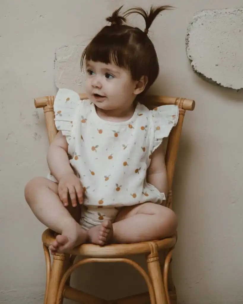 romper od muslina sa karnerom na ramenima krem boje sa motivom pomorandze na devojcici koja sedi na drvenoj stolici