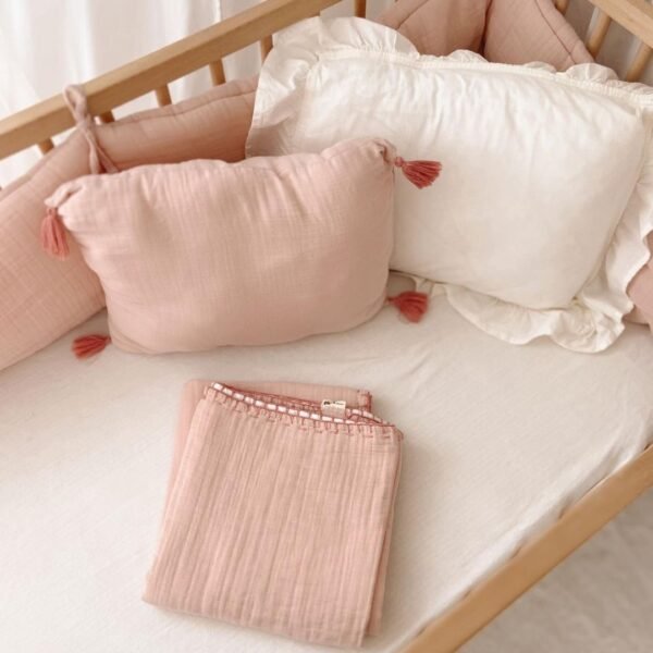 ogradica za bebe od muslina puderroze boje sa jastukom sa kicankama u kombinacijij sa belom posteljinom na krevecu prikazana izbliza